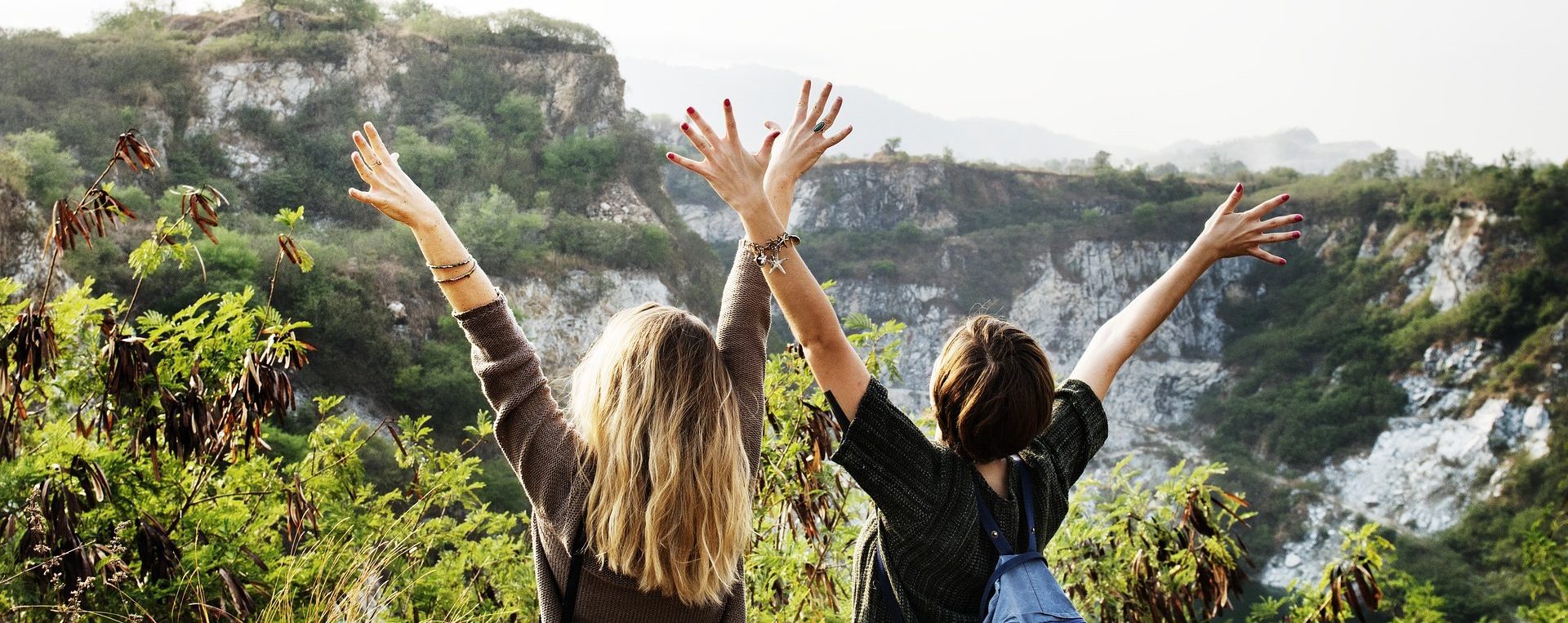 Zwei junge Frauen strecken die Arme in die Höhe und blicken auf das Gebirge vor ihnen.